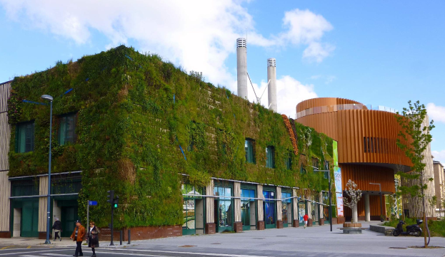 Ein großes Gebäude mit einer grünen Wand an seiner Seite, umgeben von Straßenmasten, Straßenlaternen, Verkehrspfosten, Verkehrssignalen, Kraftfahrzeugen, Fußgängern, Bäumen und einem bewölkten Himmel, als Teil des Green Building Projects.