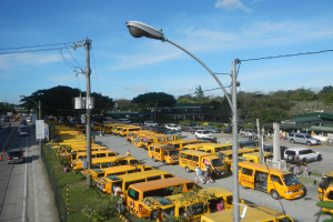 Eine große Anzahl gelber Schulbusse auf der Seite einer Straße geparkt, mit Menschen auf dem Gehweg, Strommasten mit Drähten, Laternen, Bäume, Gebäude und ein bewölkter Himmel im Hintergrund.