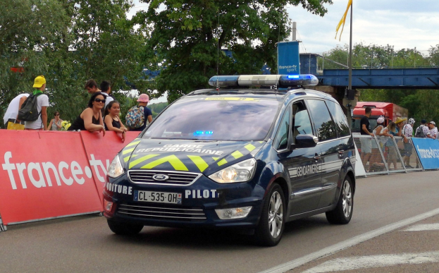 Polizeifahrzeug fährt an einer Menschenmenge vorbei, die Schilder trägt, eine Brücke, Bäume, eine Fahne und einen bewölkten Himmel im Hintergrund.