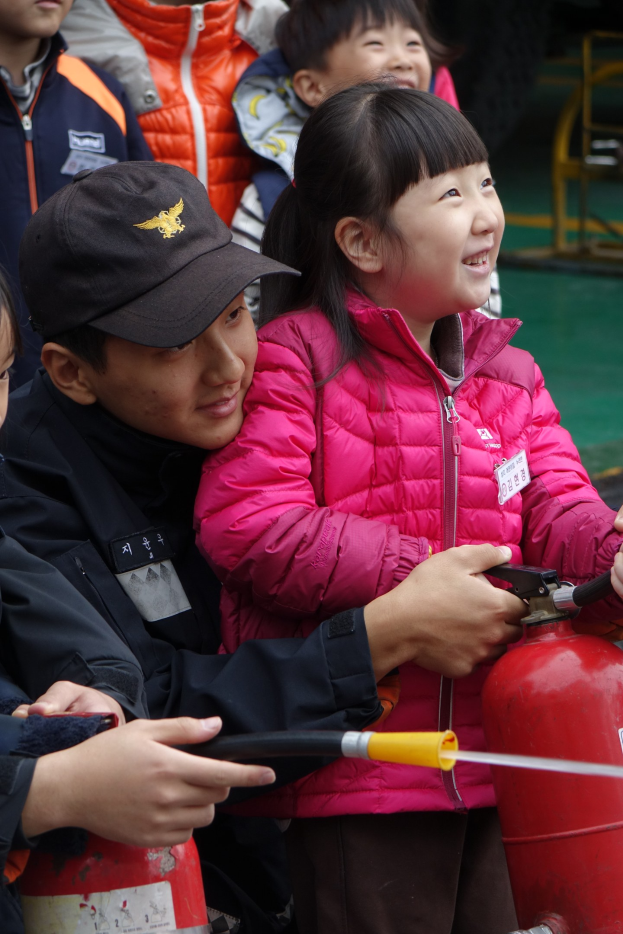 Kinder in einer Schulumgebung um ein rotes Feuerhydrant versammelt, einer hält eine Leitung, mit verschiedenen Gegenständen im Hintergrund.