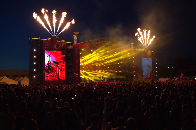 Große Menschenmenge auf einem Musikfestival mit beleuchteter Bühne mit Performern, Bannern, Zelten links und Feuerwerk am dunklen Himmel.