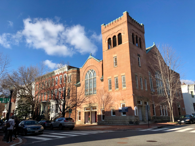 Ein großes Backsteingebäude mit einem Glockenturm, identifiziert als St. Mary's Episcopal Church, umgeben von Bäumen, Straßenlaternen, Straßenlaternen, Kraftfahrzeugen, Fußgängern und Namensschildern, unter einem bewölkten Himmel.