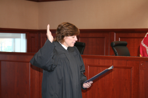 Eine Frau in einer schwarzen Robe steht an einem Richtertisch und hält eine Akte in der Hand, mit einem Mikrofon und einer Flagge auf dem Tisch, Stühle dahinter und ein Fenster in der Wand.