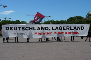 Eine Gruppe von Menschen hält ein Transparent mit der Aufschrift 'Deutschland, Lagerland Migration ist kein Verbrechen' und eine Flagge, mit Straßeninfrastruktur und Gebäuden im Hintergrund.