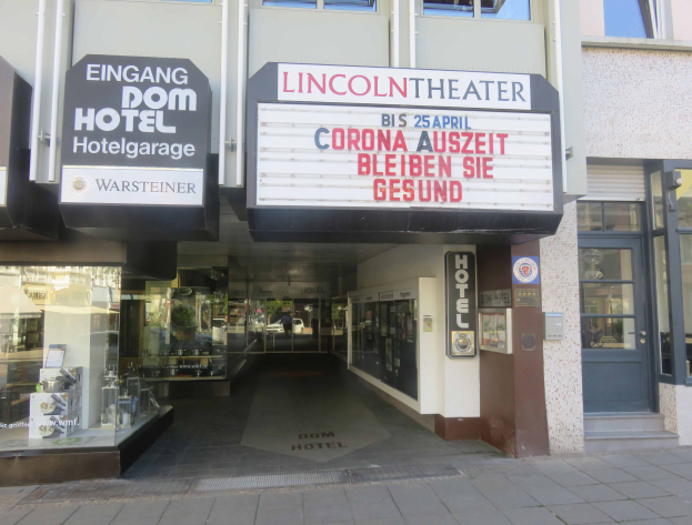 Außenansicht des Lincoln Theaters in Berlin, Deutschland, mit Glasfenstern und -türen sowie einer Tafel mit Text und einem durch das Glas sichtbaren Innenraum, der eine belebte Stadtlandschaft zeigt.