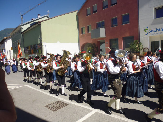 Eine Gruppe von Menschen in traditioneller bayrischer Tracht, die auf einer Straße mit Gebäuden, einigen Fahnen haltenden Personen und einem Hügel sowie einem blauen Himmel spielen.