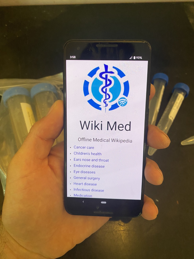 Eine Person hält ein Smartphone in der Hand, auf dem die WikiMed-App geöffnet ist, mit Reagenzgläsern und anderen Gegenständen auf dem Tisch vor ihnen.