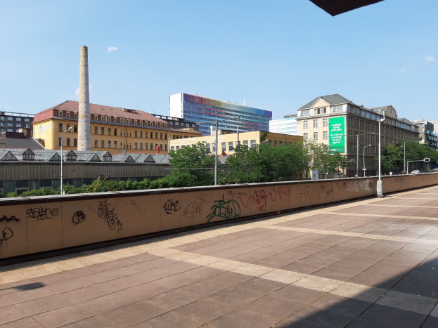 Stadtstraße mit Graffiti-beschmierten Wand, Bäumen, Straßenlaternen, Schild, Fahrzeugen, Gebäuden mit Fenstern, einem Schornstein und einem bewölkten Himmel.