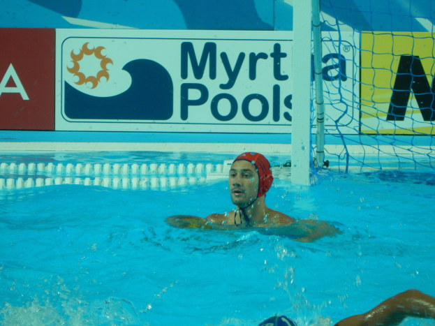 Ein Wasserballspieler mit Mütze schwimmt in einem Pool mit einem Netz auf der rechten Seite und einer Werbetafel im Hintergrund.