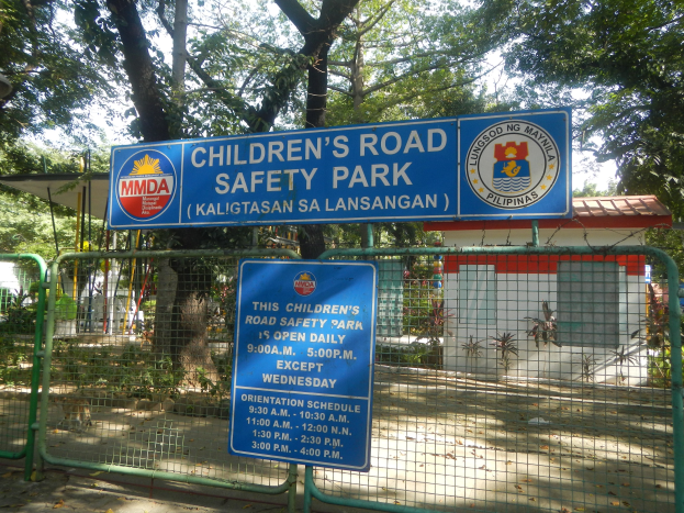 Metallzaun um einen Kinderstraßenverkehrspark mit zwei blauen Informationsschildern, einem kleinen Haus, Pflanzen, Bäumen und einem bewölkten Himmel.