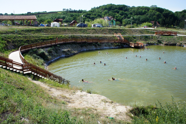 Gruppe von Menschen, die in einem Gewässer schwimmen, mit einer Brücke, Treppen, Schuppen, Fahrzeugen, Pfählen und einem klaren blauen Himmel im Hintergrund, umgeben von grünem Gras, Pflanzen und Bäumen.