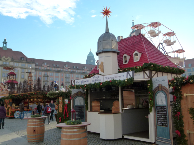 Ein belebter Weihnachtsmarkt in Nürnberg, Deutschland mit Menschen um geschmückte Stände, festliche Lichter, ein Riesenrad, Gebäude und eine Tafel mit Text auf der rechten Seite.