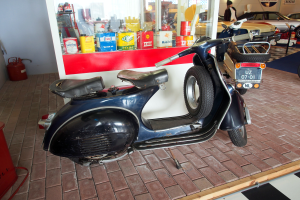 Ein schwarzer und weißer Roller steht vor einer Tankstelle, mit einem roten Objekt und einem Feuerlöscher auf der linken Seite und mehreren Fahrzeugen, einer Person und Brettern im Hintergrund.
