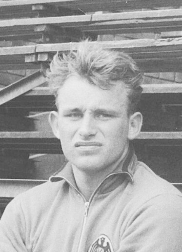 Schwarzes und weißes Foto eines Mannes in einer Jacke, der vor gestapelten Holzplanken steht, der als legendärer deutscher Fußballspieler gilt.