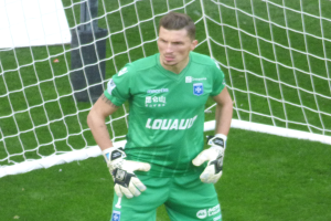 Ein Fussballtorwart in grüner Uniform steht vor einem Tor auf grünem Rasen und trägt Handschuhe.