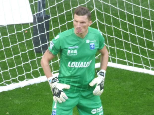 Ein Fussballtorwart in grüner Uniform steht vor einem Tor auf grünem Rasen und trägt Handschuhe.
