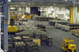 Großes Lagerinnere mit mehreren Lastwagen, Shipping-Containern, Menschen, Deckenbeleuchtung, Metallstangen und einem sichtbaren Rad.