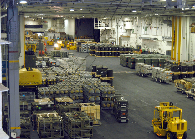 Großes Lagerinnere mit mehreren Lastwagen, Shipping-Containern, Menschen, Deckenbeleuchtung, Metallstangen und einem sichtbaren Rad.