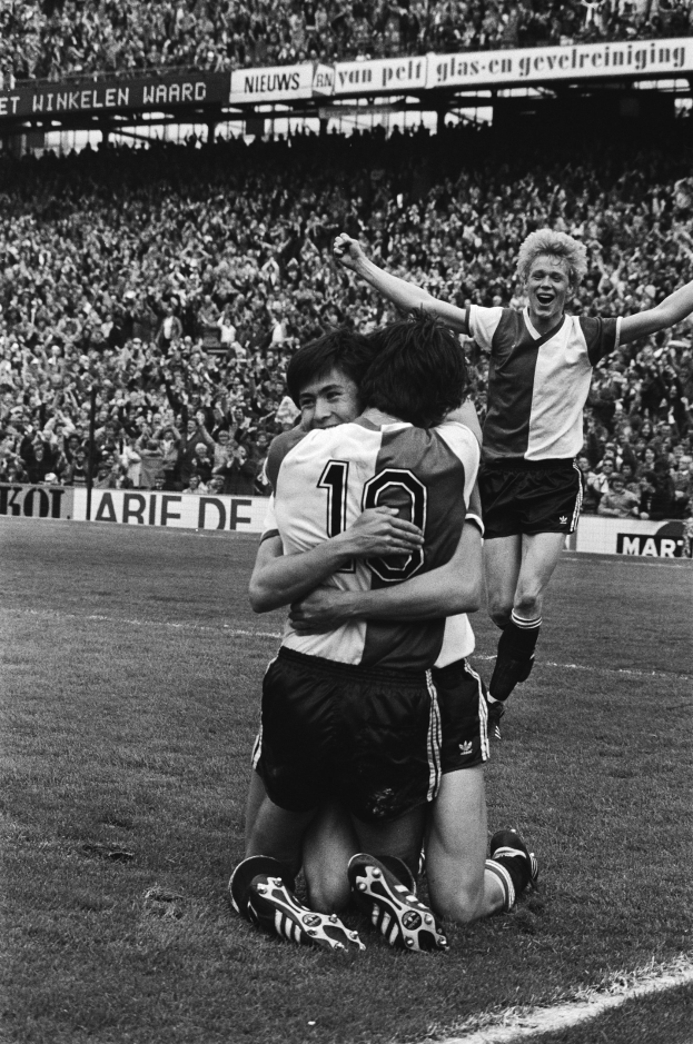 Drei Fußballspieler feiern ein Tor auf dem Boden, mit jubelnder Menge und schwenkenden Fahnen im Hintergrund, in Schwarz-Weiß.