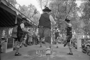Eine Gruppe von Männern in traditioneller bayischer Tracht tanzt auf einer Bühne vor einem gemischten Publikum, mit Bäumen und einer Hütte im Hintergrund, dargestellt in Schwarz-Weiß.