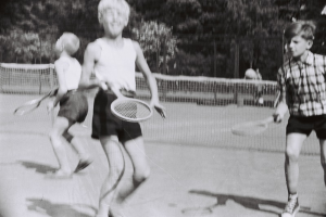 Drei Kinder beim Tennis auf einem Platz, jeder hält einen Schläger, mit einem Netz, einigen Menschen und Bäumen im Hintergrund.