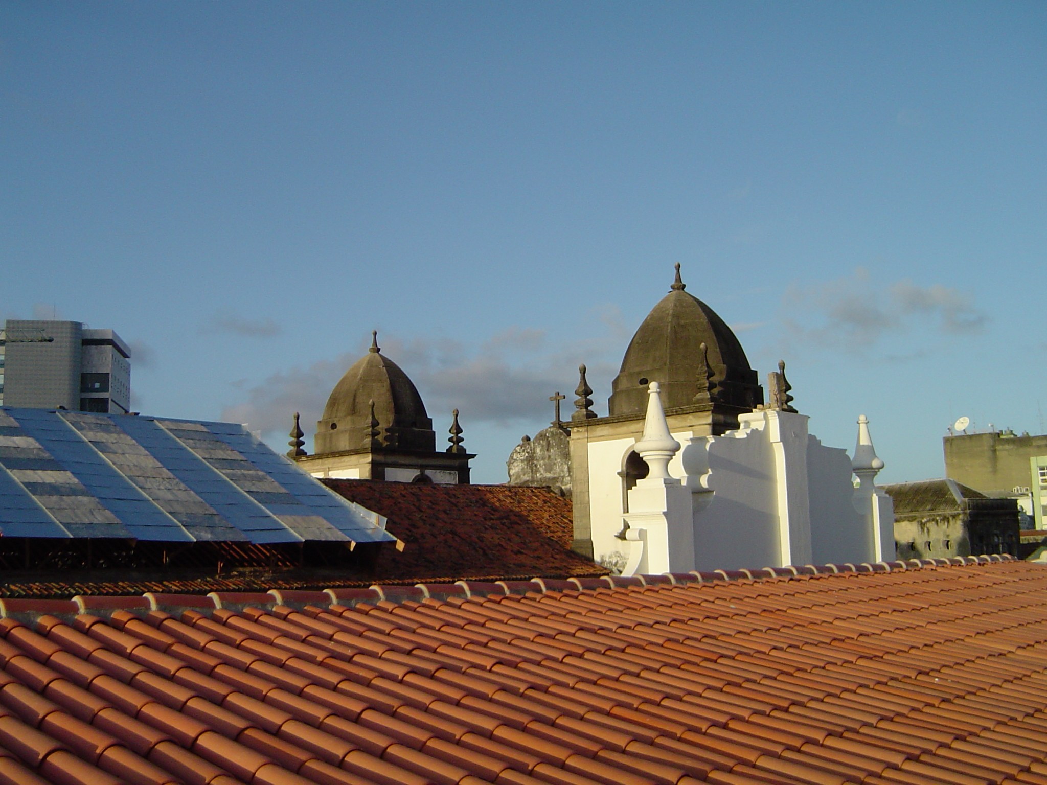 Stadtansicht mit Gebäuden im Vordergrund und einem blauen Himmel im Hintergrund, mit Solarpanels auf einem Dach, die den Einsatz erneuerbarer Energien anzeigen.