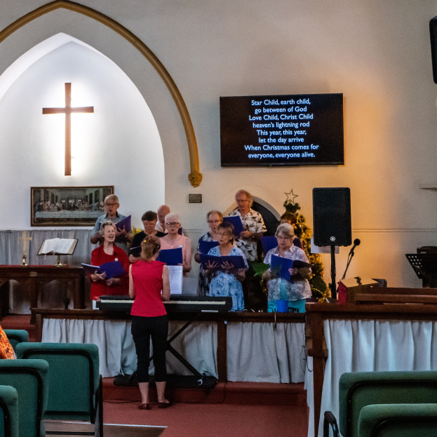Eine Gruppe von Menschen steht vor einer Kirche, einige halten Bücher, Stühle links, Kreuzsymbol und Foto Rahmen an der Wand, ein Schild mit Text, Lautsprecher mit Ständer und ein Tisch mit einer Decke, scheinbar singend als Chor.