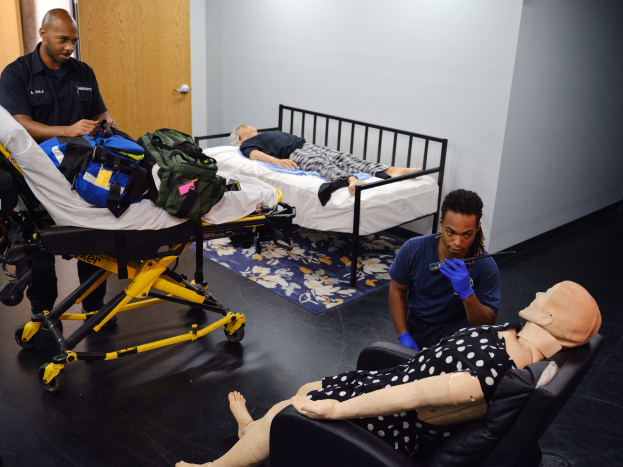 Eine Erste-Hilfe-Ausbildungsanlage mit zwei Personen auf Betten, einer Person stehend mit einem Gegenstand in der Hand, einer weiteren Person sitzend auf einem Stuhl, einer Puppe auf einer Trage, einer Tasche auf der Trage und einer Matte auf dem Boden.