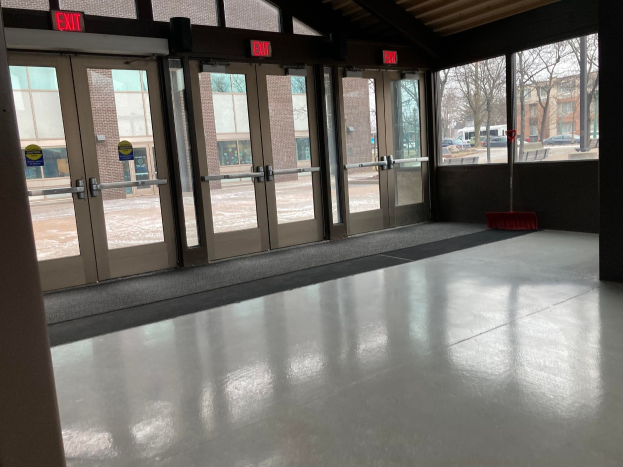 Großer Raum mit mehreren Fenstern und Türen, Besenstiel auf dem Boden, Schilder, Dach, Bäume im Freien, Fahrzeuge auf dem Boden, nahe Gebäude und bewölkter Himmel, was auf einen gewerblichen Bodenreinigungsservice hinweist.