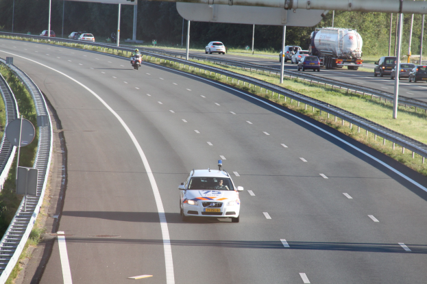 Ein Auto auf einer Autobahn mit einem stationären Polizeiwagen am Rande, andere Fahrzeuge und Infrastruktur wie Gelönder, Masten, Schilder und Bäume im Hintergrund.
