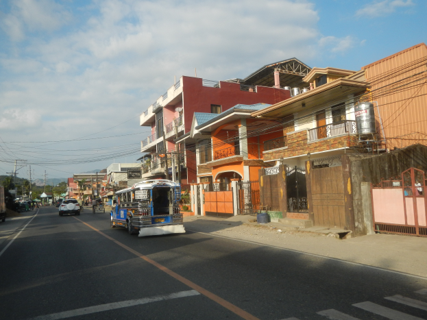 Eine Straße in einer Stadt mit einem Bus, der daran vorbeifährt, umgeben von hohen Gebäuden mit Fenstern, Geländern und Türen, Strommästen mit Kabeln, Bäumen und einem bewölktem Himmel im Hintergrund.