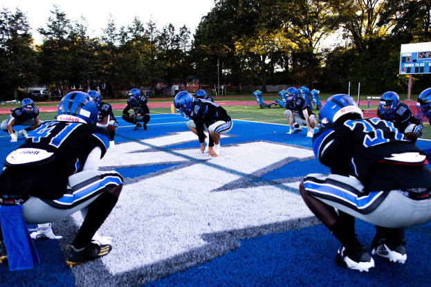 Gruppe junger Männer in Sportkleidung und Helmen, die Football auf einem Feld mit einem Scoreboard und Bäumen im Hintergrund unter einem klaren blauen Himmel spielen.