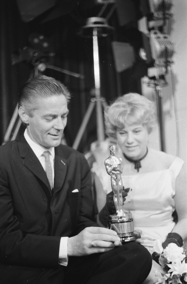 Ein Schwarz-Wei├č-Bild eines formell gekleideten Mannes mit einer Oscar-Statue und einer Frau mit einem Blumenstra├čus, die nebeneinander sitzen, mit beleuchtetem Hintergrund.