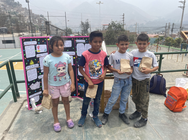 Gruppe von Kindern auf einem Basketballfeld, die Papierbeutel halten, mit Beuteln und Gegenständen rechts und einer Tafel mit Text links, vor dem Hintergrund von Zaun, Pfosten, Straßenlaternen, Bäumen, Gebäuden, Hügeln und Himmel.