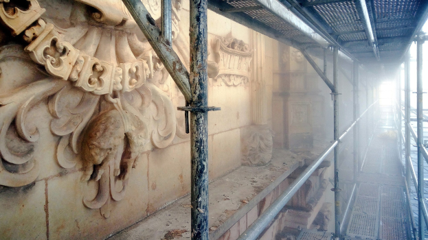 Ein renoviertes Gebäude mit detaillierten Wandverzierungen auf der linken Seite, Metallstangen auf der rechten Seite und Gerüsten drumherum.