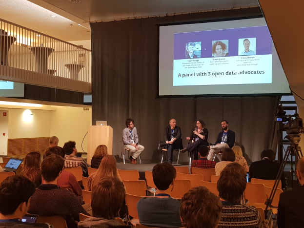 Eine Gruppe von Menschen, die auf Stühlen vor einem Projektionsschirm sitzen, mit einem Laptop auf einem Tisch, einer Kamera auf einem Stativ rechts daneben und Geländern, Lampen und einer Wand im Hintergrund, die an einer Podiumsdiskussion über Open-Data-Aktivisten teilnehmen.