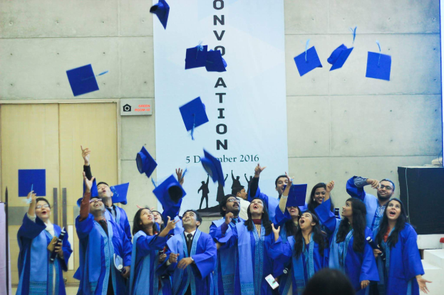 Eine Gruppe von Menschen in blauen Abschlussroben und -hüten wirft ihre Hüte in die Luft, während im Hintergrund ein Banner, eine Wand, eine Tür, ein Lautsprecherkasten und andere Gegenstände zu sehen sind und weitere Personen unten wahrscheinlich die Universität Hyderabad feiern.