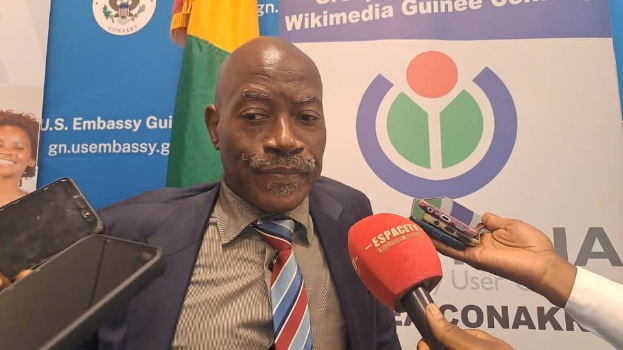 Ein Mann im Anzug spricht mit Reportern, während eine Person eine Hand mit einem Mikrofon und einem Gerät im Vordergrund hält. Im Hintergrund sind Banner, Flaggen und eine Frau zu sehen.