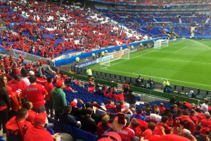 Ein volles Stadion mit sitzenden und stehenden Zuschauern, die ein Fußballspiel beobachten, mit Bannern, einem Torpfosten und Deckenbeleuchtung.