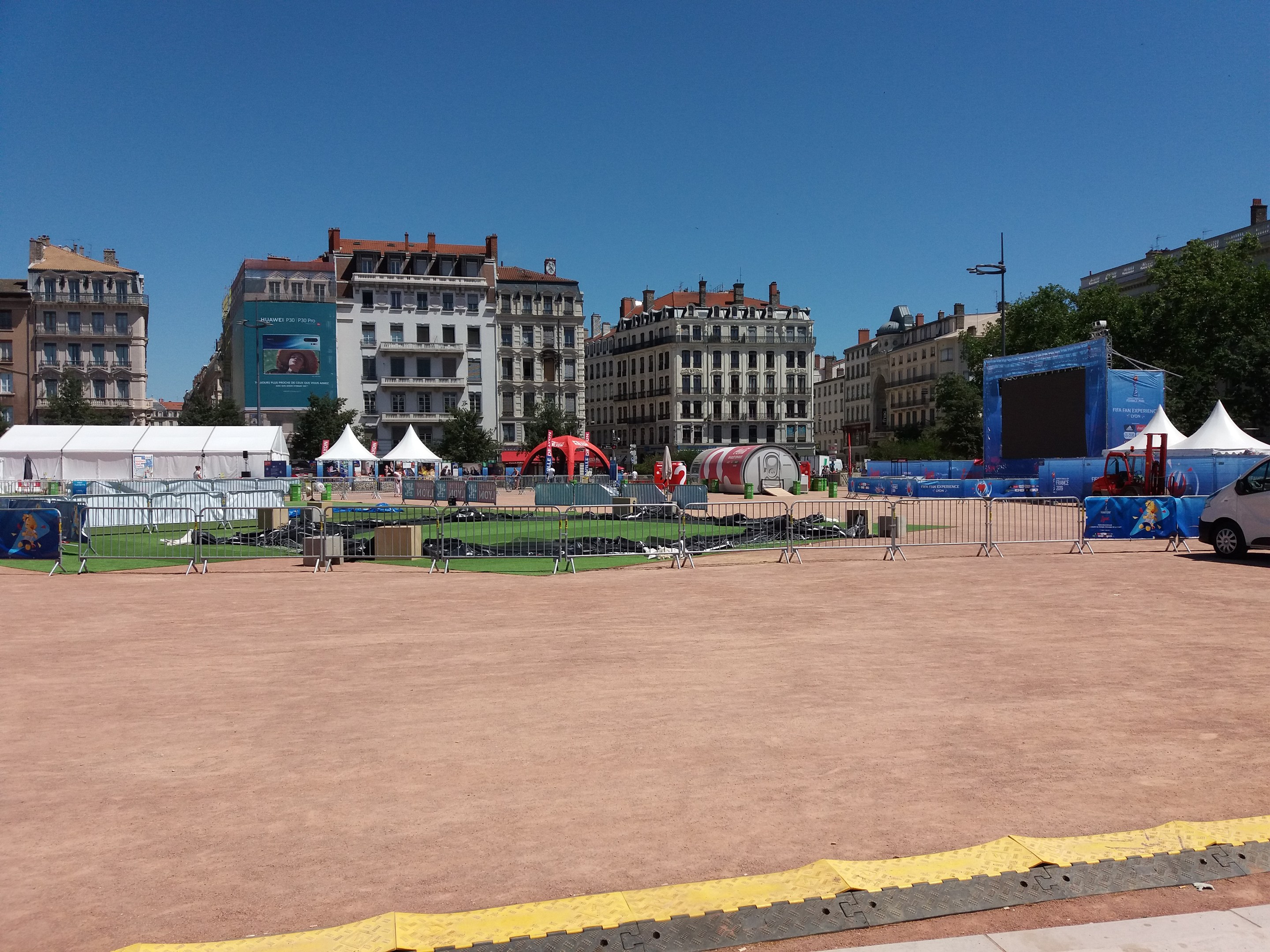 Großer offener Platz voller Numerous Zelte, umgeben von Gebäuden mit Fenstern, Bäumen und einem klaren blauen Himmel, mit Fahrzeugen auf der Straße und einem umliegenden Geländer.