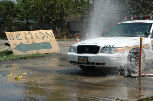 Ein Polizeiwagen fährt durch eine Pfütze, mit einer Tafel, Verkehrskegel und anderen Gegenständen am Boden; Trees, Gebäude und ein klarer blauer Himmel sind im Hintergrund zu sehen.