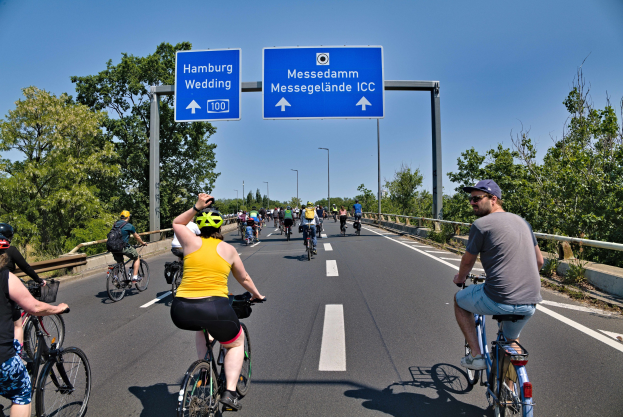 Eine Gruppe von Radfahrern in Helmen fährt auf einer Straße mit einer Begrenzung auf einer Seite und Bäumen auf der anderen, unter einem klaren blauen Himmel mit Laternen im Hintergrund; ein Schild oben zeigt eine Radtour in Hamburg.