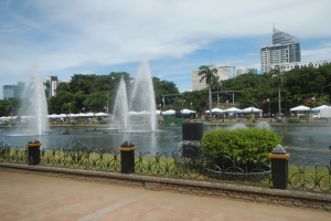 Ein Brunnen in einem Park umgeben von einem Zaun, Gras und Bäumen, mit Menschen, Fahrzeugen, Zelten, Fahnen und Gebäuden im Hintergrund unter einem bewölkten Himmel.