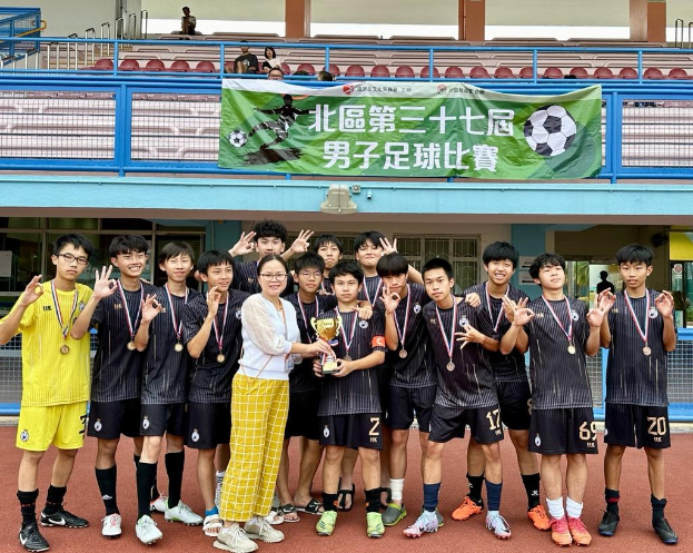 Eine Gruppe junger Männer in Fußballtrikots steht auf einem Feld, hält einen Pokal und trägt Medaillen, mit einem Gebäude im Hintergrund, auf dem 'Yokohama U-16 Jungenfußballmannschaft' steht, und Zuschauern in der Nähe.