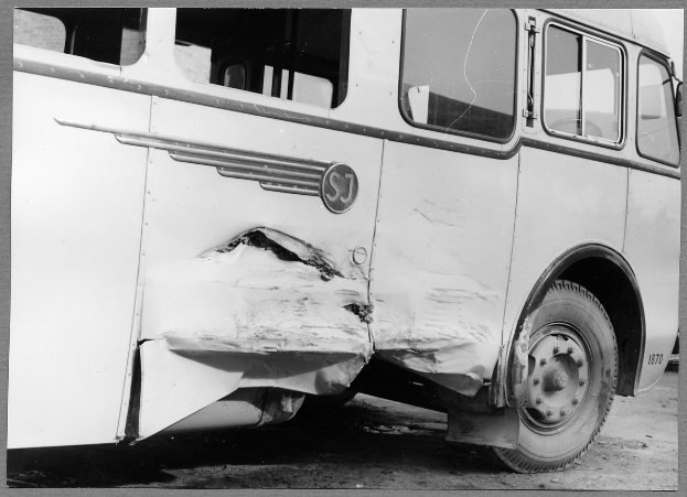 Ein Schwarz-Weiß-Foto eines beschädigten Busses am Straßenrand, dessen Vorderseite eingedrückt ist, Fenster zerbrochen und die Fahrertür offen steht.