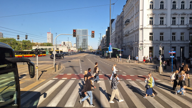 Eine Gruppe von Menschen überquert eine Straße an einer Ampel mit einem Bus auf der linken Seite, Fahrzeugen auf der Straße, Verkehrszeichen, Laternenmasten, Gebäuden, Bäumen und einem klaren blauen Himmel.