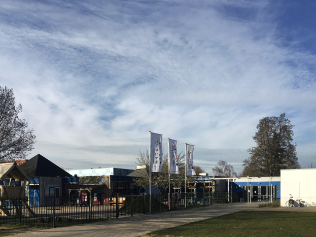 Großes Gebäude mit einem grasigen Bereich davor, umgeben von einem Metallzaun, Bäumen, Bannern, einem Fahrrad, nahen Häusern und einem bewölkten Himmel, identifiziert als der neue Spielbereich für Kinder an der Schule.