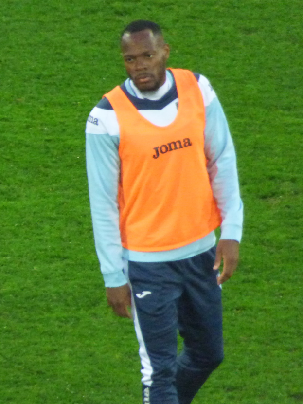 Ein Footballspieler in einem orangen T-Shirt und blauen Hosen läuft entschlossen über ein grünes Feld.