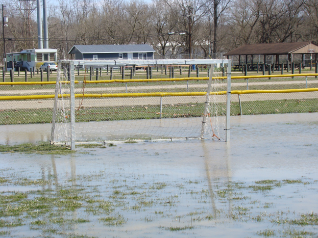 Ein Fußballtor steht auf einem überfluteten Feld mit Gras und Wasser, umgeben von Hütten, Pfählen, Bäumen, Fahrzeugen und einem klaren blauen Himmel im Hintergrund.