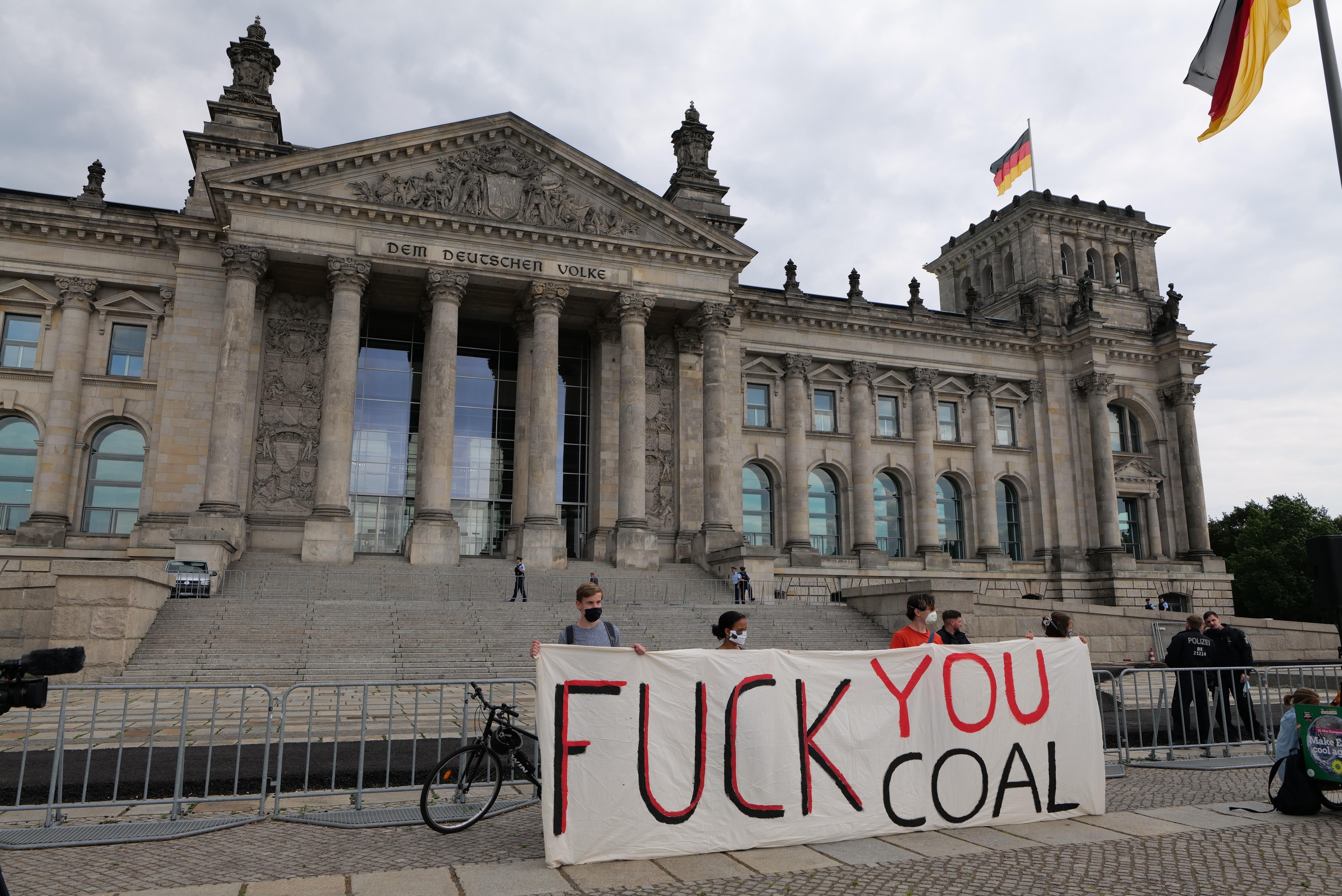Protestierende mit einem "Fuck You Coal"-Schild vor dem Reichstaggebäude in Berlin, Deutschland, mit Architektur des Gebäudes, einem Fahrrad, Bäumen, einer Fahnenstange und bewölktem Himmel.